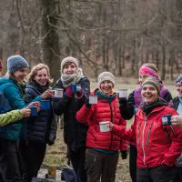 Eine Gruppe von Menschen in Winterkleidung steht draußen in einem Wald, hält Metallbecher und lächelt sich an. Die Personen sind mit bunten Jacken, Mützen und Schals bekleidet. Im Hintergrund sind blattlose Bäume und weiches, diffuses Licht zu sehen.