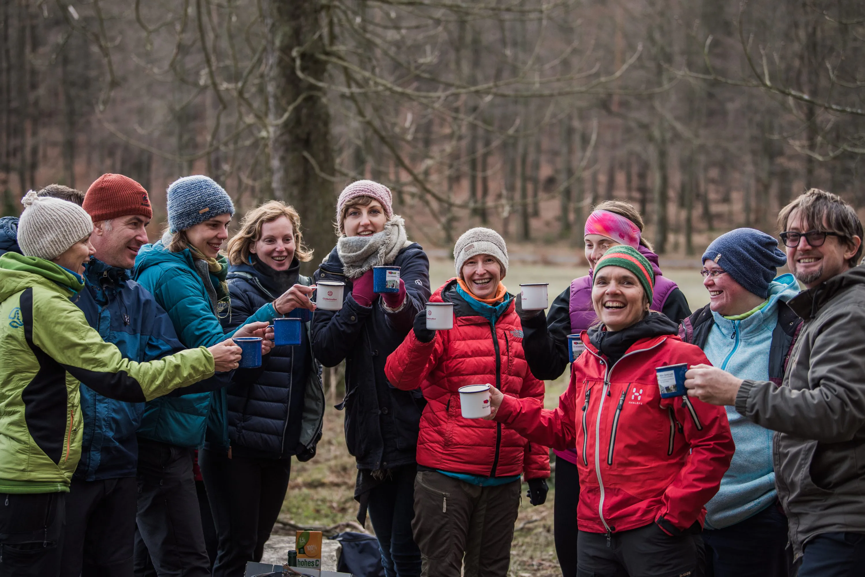 Eine Gruppe von Menschen in Winterkleidung steht draußen in einem Wald, hält Metallbecher und lächelt sich an. Die Personen sind mit bunten Jacken, Mützen und Schals bekleidet. Im Hintergrund sind blattlose Bäume und weiches, diffuses Licht zu sehen.