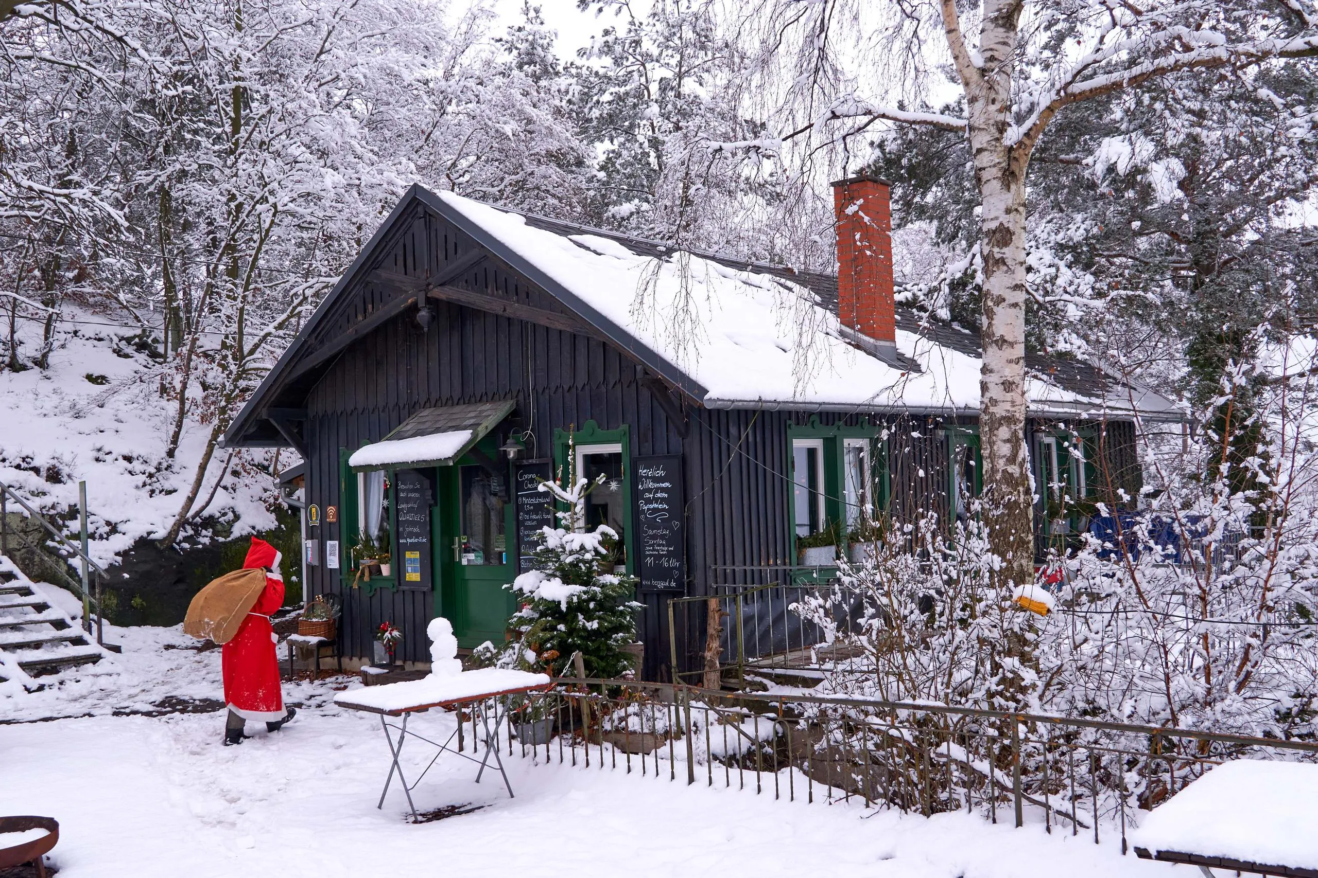 Eine als Weihnachtsmann verkleidete Person, die einen großen Sack trägt, geht in einem Wald auf eine schneebedeckte Holzhütte zu, an der Menüs auf einer Kreidetafel stehen.