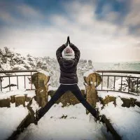 Person steht mit erhobenen Armen und weit gespreizten Beinen auf einer verschneiten Aussichtsplattform und scheint eine Yoga-Pose einzunehmen. Der Hintergrund zeigt eine malerische, schneebedeckte Berglandschaft und einen bewölkten Himmel.