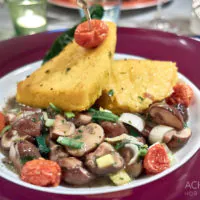 Ein Gourmetgericht bestehend aus zwei großen Polentascheiben, belegt mit gerösteten Kirschtomaten auf einem Bett aus sautierten Pilzen und gehackten Frühlingszwiebeln, serviert auf einem weißen Teller mit kastanienbraunem Rand. Der Teller steht auf einem Tisch mit Kerzenlichtatmosphäre. .