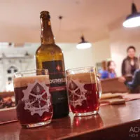 Auf einem Holztisch steht eine Flasche Dunkles-Bier neben zwei gefüllten Biergläsern, die jeweils ein Logo und „Seit 2005“ tragen. Im Hintergrund sieht man Menschen in einem warm beleuchteten Raum mit Hängelampen. Der Bildnachweis lautet „ACHIMEURER HDR PHOTOGRAPHY“ in der unteren rechten Ecke.