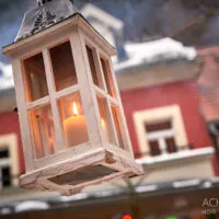 Eine weiße Laterne mit einer brennenden Kerze darin hängt im Freien vor einem Hintergrund aus schneebedeckten Gebäuden. Die Gebäude haben unterschiedliche Farben und architektonische Details. Im Hintergrund hängen einige Lichter, die für eine festliche Atmosphäre sorgen. Der Bildnachweis lautet „ACHIMEURER HDR PHOTOGRAPHY“.