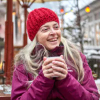 Eine Frau mit langen, blonden Haaren lächelt und hält in beiden Händen ein warmes Getränk. Sie trägt eine rote Strickmütze und eine magentafarbene Jacke. An einem verschneiten Tag sitzt sie draußen an einem Holztisch mit Gebäuden und festlichen Lichtern im Hintergrund.
