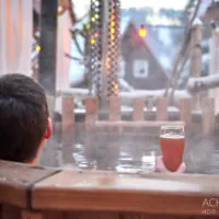 Zwei Personen entspannen in einem hölzernen Whirlpool im Freien. Eine Person hält ein Glas Bier. Dampf steigt aus dem Whirlpool auf, im Hintergrund leuchten Lichterketten und eine verschneite Landschaft. Die Atmosphäre wirkt gemütlich und heiter.