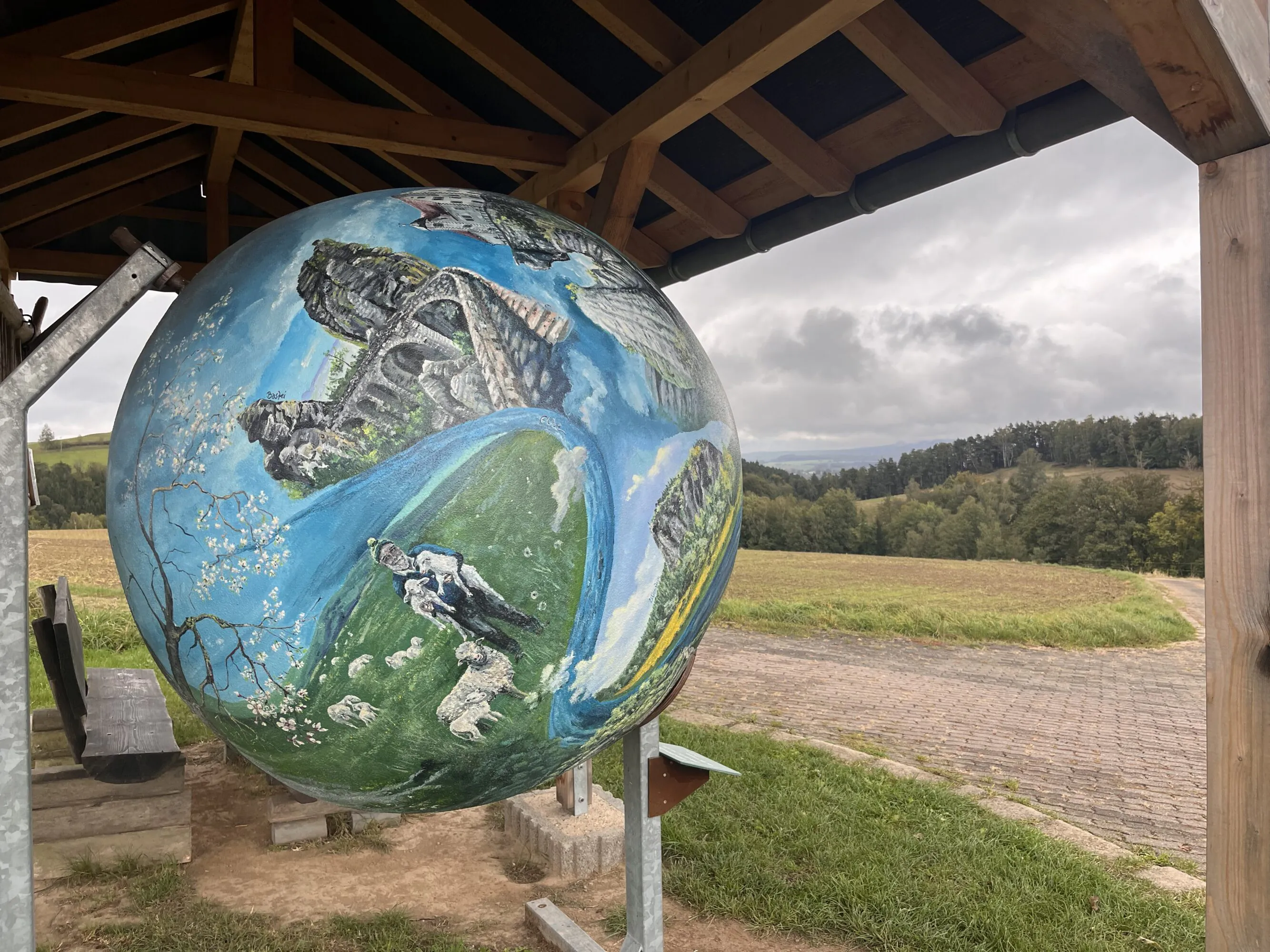 Eine große kugelförmige Skulptur, bemalt mit einer Landschaftsszene, in der ein Hirte, Schafe und Felsformationen zu sehen sind, ausgestellt unter einem hölzernen Unterstand mit Blick auf ein ländliches Feld.