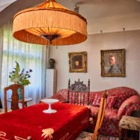 Ein Wohnzimmer im Vintage-Stil mit einer roten Tischdecke, antiken Stühlen, einem Sofa mit Blumenmuster, einer großen Lampe mit Fransen und mehreren klassischen Porträts an den Wänden. Sonnenlicht fällt durch die transparenten Vorhänge hinter den Möbeln.