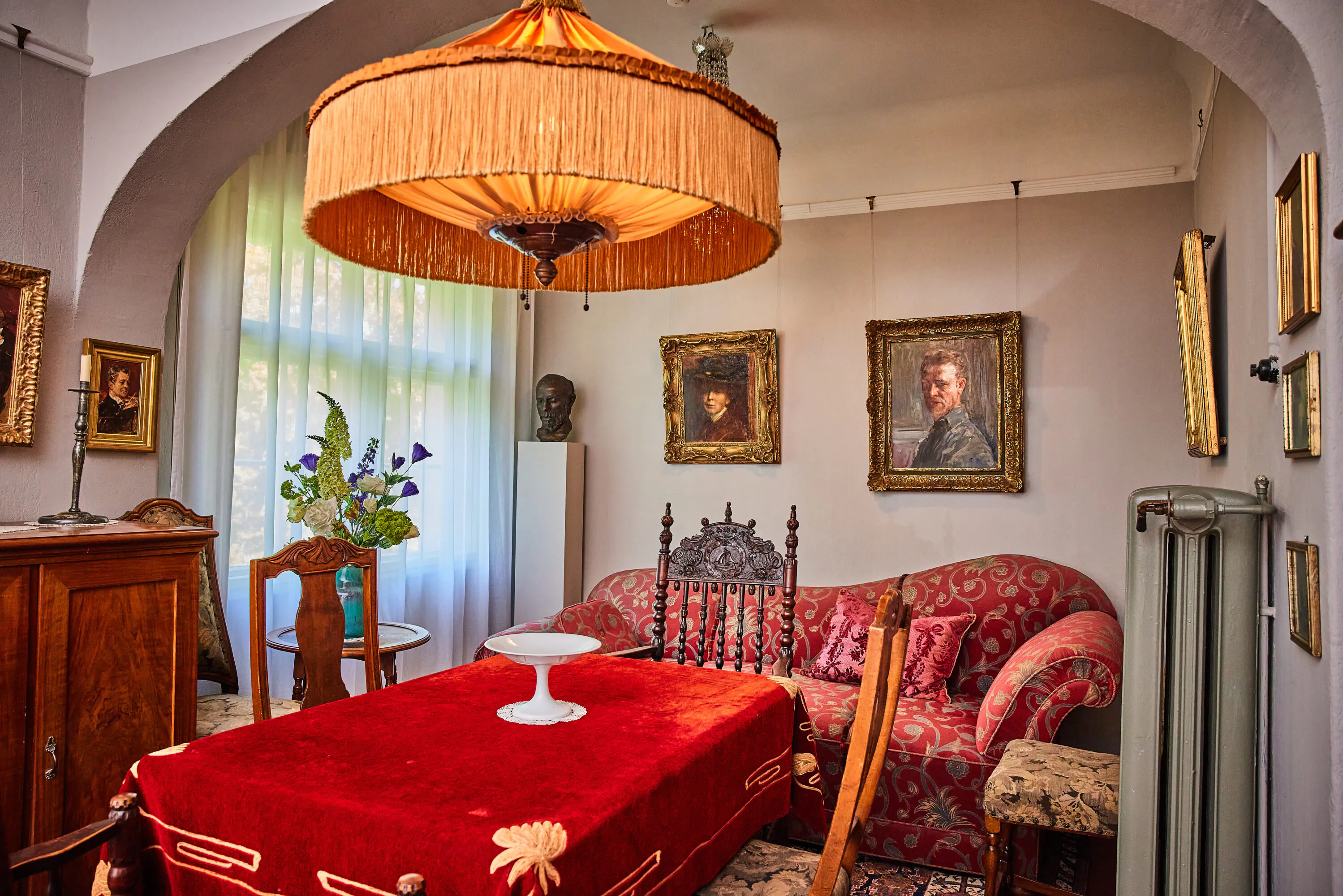 Ein Wohnzimmer im Vintage-Stil mit einer roten Tischdecke, antiken Stühlen, einem Sofa mit Blumenmuster, einer großen Lampe mit Fransen und mehreren klassischen Porträts an den Wänden. Sonnenlicht fällt durch die transparenten Vorhänge hinter den Möbeln.