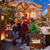 Drei warm angezogene Personen sitzen an einem Tisch im Freien neben einer Feuerstelle und schlürfen heiße Getränke. Schneebedeckte Bäume und festliche Lichter schmücken den Hintergrund in einer gemütlichen Winteratmosphäre. Ein großer Sonnenschirm bietet Schutz über dem Kopf.
