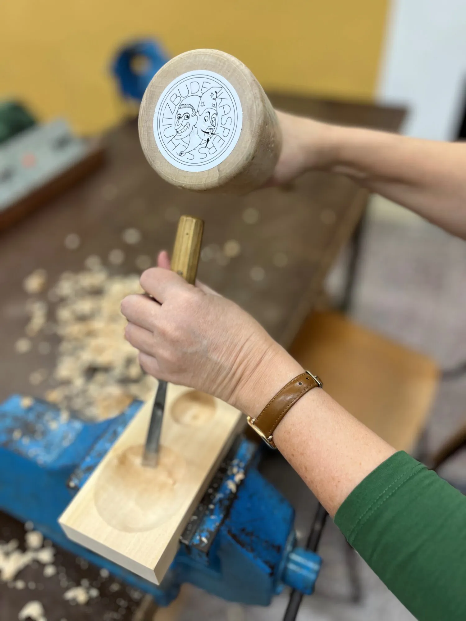 Eine Person, die mit einem Meißel und einem Holzhammer Holz bearbeitet und dabei eine runde Rille in einen Holzblock fräst, der in einem blauen Schraubstock eingespannt ist. Auf dem Tisch liegen Holzspäne verstreut.