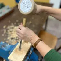 Eine Person, die mit einem Meißel und einem Holzhammer Holz bearbeitet und dabei eine runde Rille in einen Holzblock fräst, der in einem blauen Schraubstock eingespannt ist. Auf dem Tisch liegen Holzspäne verstreut.
